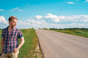 A young blond man in a checked shirt walks in the countryside with fields near the highway, copy space