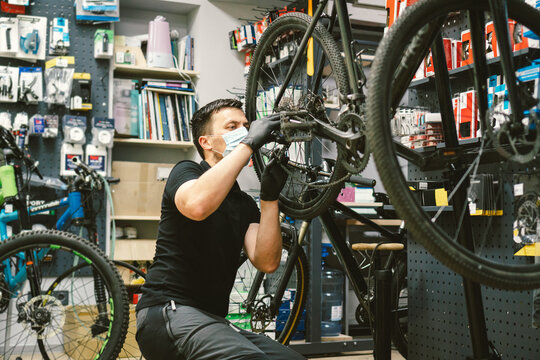 Young man working in a biking repair shop under quarantine and pandemic coronavirus in a protective mask on the face. Bicycle Mechanic Repair Bike, Cycle Workshop in medical mask covid 19
