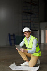 Portrait of young engineer in work helmet smiling at camera while working with blueprint and digital tablet in construction site
