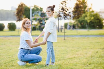 Fototapeta premium Attractive young woman with her little cute daughter are spending time together outdoors. Mom with daughter in park on a green grass during the sunset.