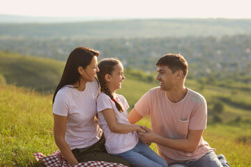 Joyful family relaxing on picnic blanket in mountains. Cute girl laughing with her parents on summer day in countryside