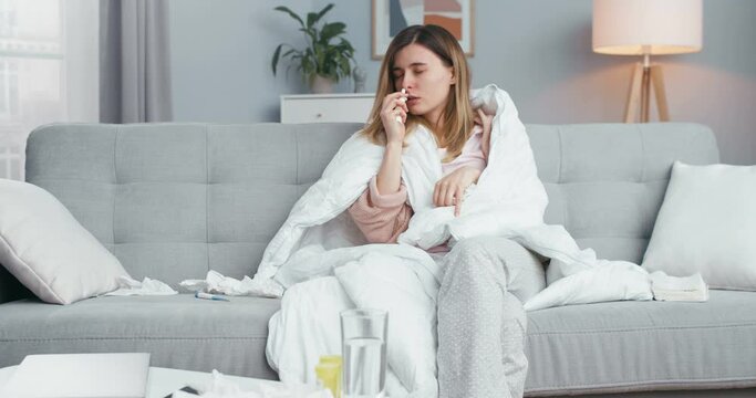 Young Caucasian ill girl with coronavirus sitting under blanket on couch in living room, spraying medical drops in nose, caughing and sneezing. Sick woman with coronavirus sympthoms.