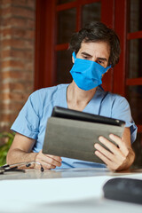 Doctor with protective mask working on a Tablet after hours at home. Work from home to avoid the concept of Coronavirus COVID 19.