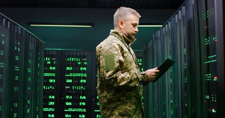 Portrait shot of Caucasian handsome man in camouflage uniform holding tablet device and looking straight at camera while walking dark passage among servers. US army officer in monitoring room.