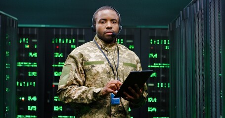 Portrait of young African American handsome army technician in uniform and headset holding tablet...