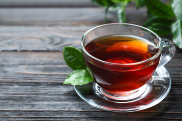 Glass cup of aromatic black tea and green leaves on wooden table, space for text