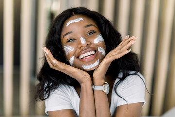 Spa, skin care, female facial treatment. Close up portrait of joyful African woman applying white mud facial mask touching her face and smiling, looking at camera. Cosy home interior