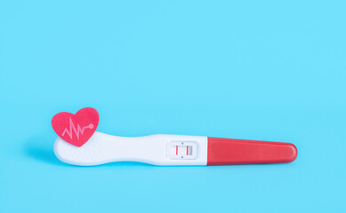 Positive pregnancy test and small red heart with pulse line on a blue background