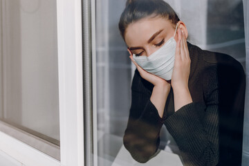 young attractive girl in a protective medical mask looks out the window. isolation during the...