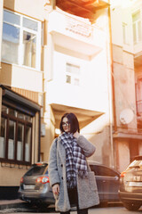 Attractive positive young girl wearing glasses in a coat on the background of buildings on cars