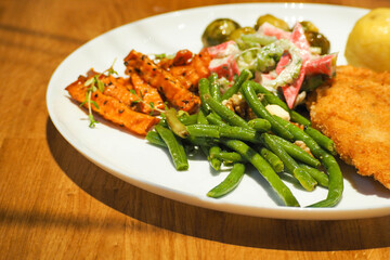 green asparagus pods and sticks of baked sweet potatoes, chop and salad with crab sticks and celery stalk on a large white round plate on a wooden table