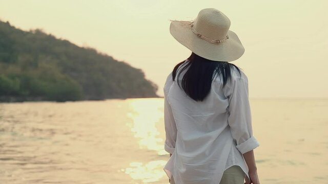 Portrait of attractive asian woman wearing fedora hat standing on the beach looking at sea view beautiful sunset. Holiday summer travel concept.