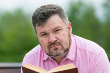 Caucasian man in a red shirt is reading a book. Adult man 40-45 years old reading fiction while sitting a park bench.