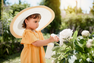 Little girl kid in yellow dress and oversize white hat spraying with water white peonies flowers.