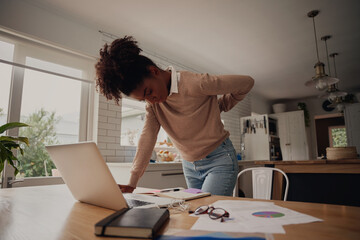 Young afro woman standing suffering from back pain while working on laptop at home