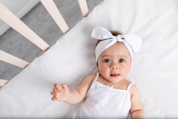 portrait of a smiling baby girl 6 months old lying in a crib in the children's room on her back with a bow on her head, looking at the camera, baby's morning, children's goods concept