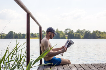 a man reading a fresh press on a wooden pier sitting by the lake