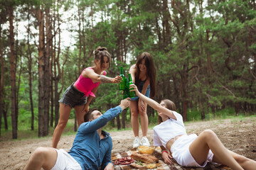 Togetherness. Group of friends clinking beer bottles during picnic in summer forest. Lifestyle, friendship, having fun, weekend and resting concept. Looks cheerful, happy, celebrating, festive.