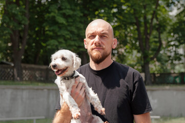 Regular serious bald man in black t-shirt holding a little white dog in his hands on the street