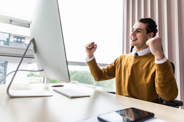 Excited young entrepreneur working with a pc and reading good news on line at workplace in office