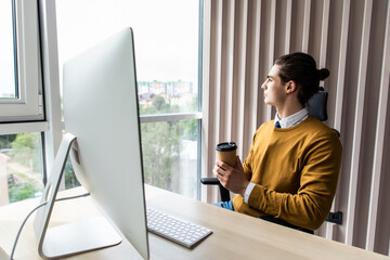 Young business man looking at pc computer screen and holding coffee cup in office