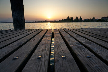 Naklejka na ściany i meble Pier wooden plank on lake during sunrise background. Wood board perspective on the Garda lake water with waves and warm colors.