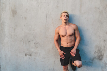 Natural portrait of young athletic shirtless man standing near concrete wall.