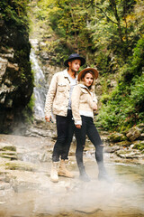 Young and beautiful couple at the mountain waterfall - Happy tourists visiting mountains. Lovestory. Tourists in hats. Military fashion.