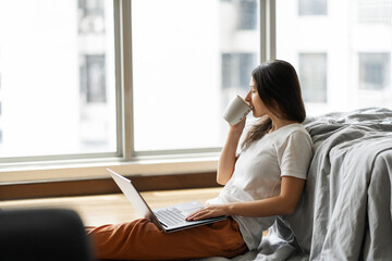 Beautiful young brunette girl working on a laptop and drinking coffee, sitting on the floor near the bed by the panoramic window. Stylish modern interior. A cozy workplace. Shopping on the Internet