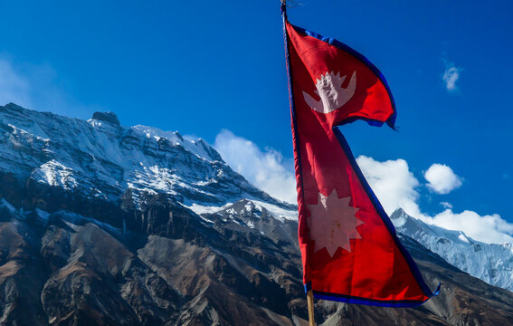 A Nepalese flag waving smoothly on the wind with the view on high, snow capped Himalayan peaks, along Annapurna Circuit Trek in Nepal. Barren and sharp slopes. Exploration and discovering new places.