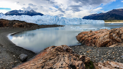 Perito Moreno Glacier reflecting in glacial lake in front of it with rocks in foreground and mountains in the background 