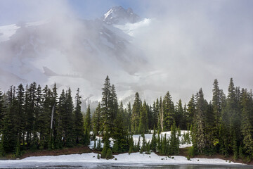 A snow covered mountain peak makes a reluctant appearance behind drifting morning clouds.  A partially thawed lake is visible in the foreground surrounded by evergreen trees.