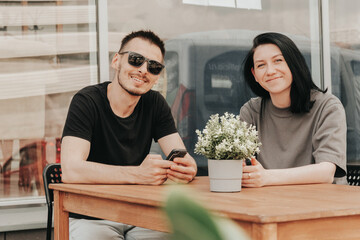 A young woman and man sits on an open veranda in a cafe and uses a smartphone while waiting for order and takeaway. 