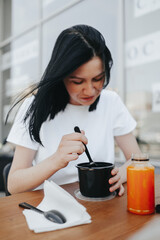 A young woman with black hair in a white T-shirt sits on an open veranda in a cafe and eats food from disposable plastic dishes. 