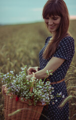 A girl in a blue dress in a white flower with a strap stands in the middle of a wheat field. In his hands he holds a wicker basket. In a straw basket a bouquet of white daisies. Around the ears 