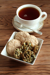 Almond cookies and sugar-coated pumpkin seeds cookies in a square bowl and a cup of black tea on a wooden table. Closeup