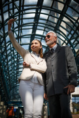 An older businessman in an elegant suit and his beautiful young associate are takeing sefie during a work break