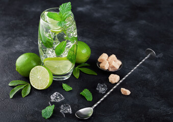 Mojito summer cocktail in glass with ice cubes mint and lime on black board with spoon and fresh...