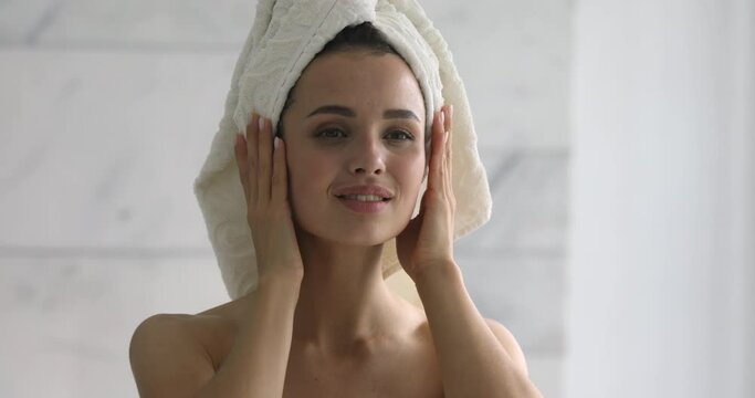 Happy attractive young woman with towel on head touch face after applying cream remove makeup look in mirror enjoy natural beauty and healthy clean soft moisturized skin care treatment, close up view