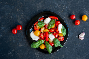 Red and yellow cherry tomato salad with pepper, basil and mozzarella on a dark background.