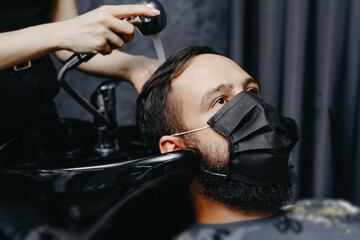 Woman barber washing hair to a bearded man in face mask. Quarantine haircut concept.