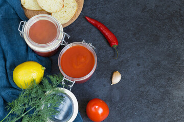 Aivar caviar in a glass jar and ingredients for it - sweet pepper, tomatoes, garlic, hot pepper, dill on a dark background. Sliced bread for snacks. Vegetarian food.