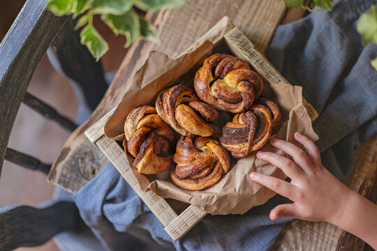 Cinnamon babka buns, homemade little babka pies in wooden box, rustic table with char, plant and child's small hand. Top view, flat lay, copy space. Kitchen with natural light, selective focus.