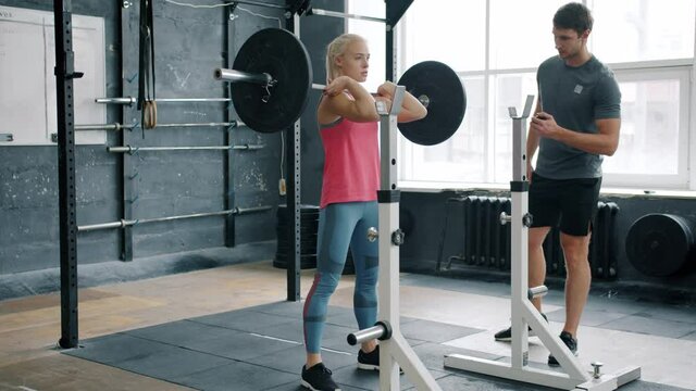 Cute blonde is squatting with barbell indoors working out with professional trainer concentrated on workout while man is using times and motivating student.