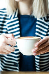 Blonde woman in blue jacket with white lines holding a cup with cappuccino. Coffee, cappuccino,...