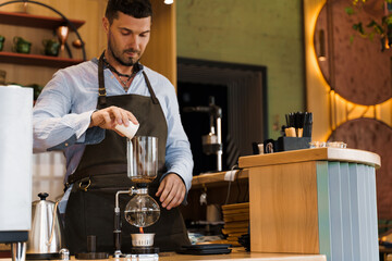 Handsome bearded barista pours freshly ground coffee to syphon device for coffee brewing in cafe. Syphon alternative method of making coffee. Scandinavian method of coffee making.