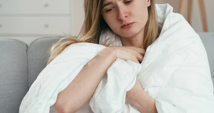 Close up of unhealthy Caucasian bad-looking young woman sitting on sofa wrapped in blanket and measuring temperature. Ill girl with flu or coronavirus having fever. Female at self-isolation.