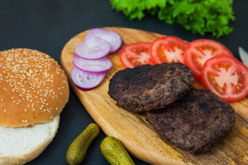 Set of beef burger ingredients on dark table. two cutlets, tomatoes, onions, salad and fresh sesame buns. Delicious homemade dish.