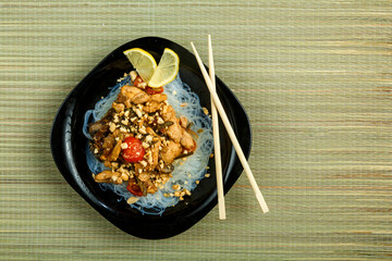 plate with funchose and chicken with vegetables in pad Thai sauce next to chopsticks on a bamboo mat.