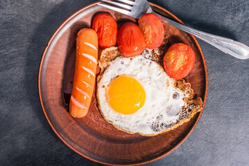 Easy quick breakfast - fried egg, tomatoes and sausage on a clay plate, top view, dark background